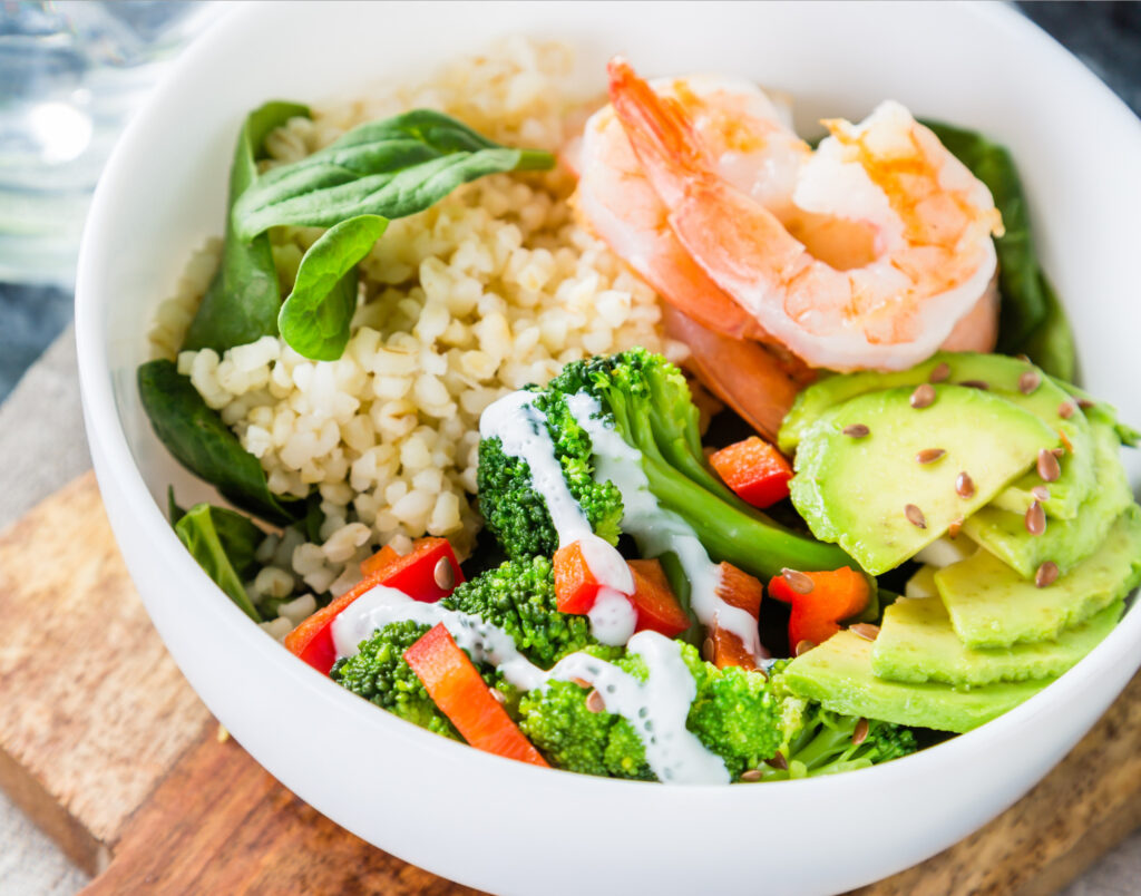 A white bowl filled with a mixture of quinoa, broccoli, shrimp, avocado, and peppers.