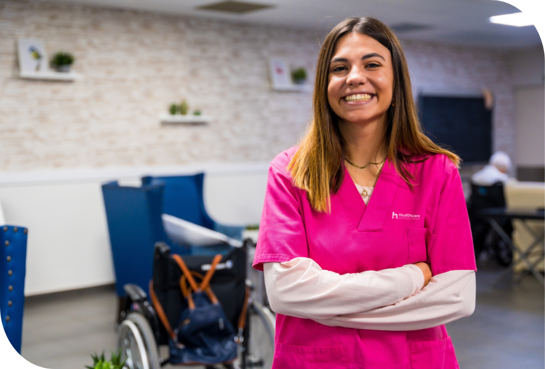 Smiling female healthcare worker wearing a pink HCSG scrub top, stands with arms crossed in a healthcare facility. Dedicated and smiling healthcare professional ready to provide quality care.